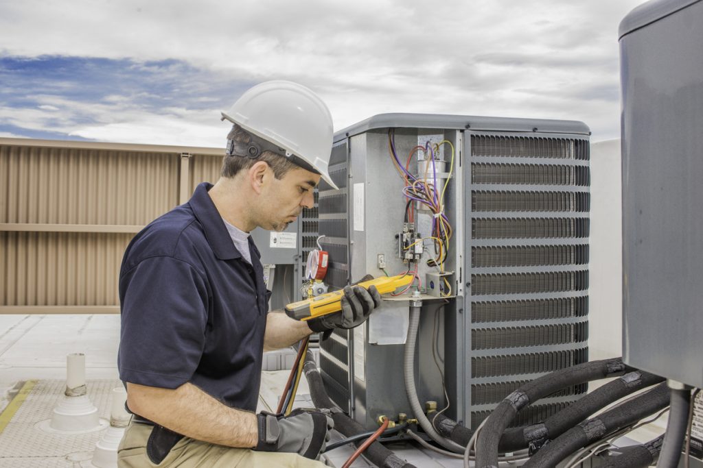 Trained hvac technician holding a voltage meter, performing preventative maintenance on a air conditioning condenser unit.